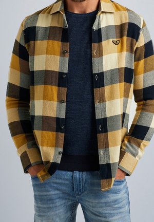Man wearing mustard, black, and cream plaid button-up shirt over a navy sweater with hands in blue jeans pockets.