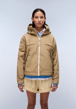 Kvinde iført en tan vindjakke med hætte, blå sweater, lyserød skjorte og opkørte beige shorts, stående mod en ensartet lys baggrund.