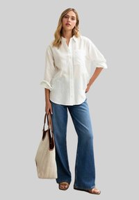 Camisa blanca a rayas con mangas remangadas, combinada con jeans azules de campana. La modelo sostiene un bolso de tela claro y lleva sandalias marrones.
