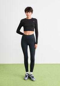 Ung kvinna står på grön golv iförd en svart kortärmad topp med lång ärm, hög midja svarta leggings, vita strumpor och silverfärgade sneakers.