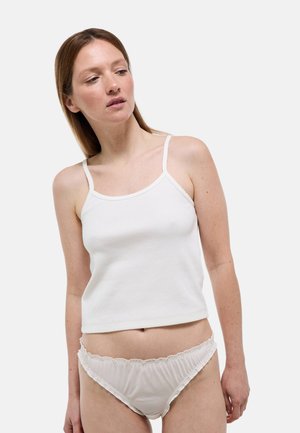 Femme portant un débardeur blanc côtelé sans manches et une culotte blanche transparente à volants, regardant légèrement vers sa gauche sur un fond uni.