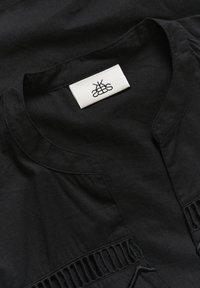 Černý oděv s kulatým výstřihem, jemným řasením po stranách a látkovou visačkou s logem uprostřed. Jemná textura.