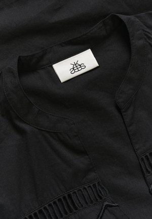 Indumento nero con scollo rotondo, pieghe sottili lungo i lati e un'etichetta in tessuto con un logo al centro. Texture morbida.