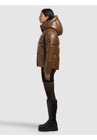 Giacca imbottita marrone con finitura lucida e cappuccio, caratterizzata da una toppa rotonda con logo nero sul braccio sinistro e tasche laterali.