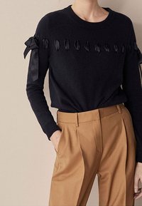 Musta neulottu neulepaita pyöreällä pääntiellä, jossa satiininauhakoristeet harteilla ja koristeellisia lehtimäisiä yksityiskohtia. Housut ovat beiget.