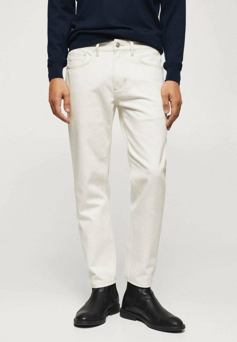 Homme portant un jean droit écru, des bottines en cuir noir et un haut sombre à manches longues, debout sur un fond clair uni.