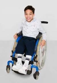 Barn i en blå rullestol, iført en lys stripet skjorte med flerfargede prikker, marineblå bukser og hvite joggesko med røde detaljer.