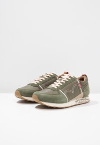 Chaussures de sport vertes composées d'un mélange de daim et de matériaux synthétiques, avec un bout rond, des lacets beige et une étiquette imprimée sur la semelle.