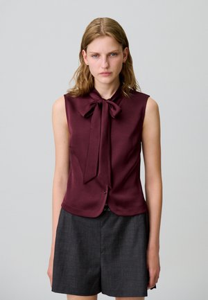 Blouse sans manches bordeaux avec un col à nœud, en tissu lisse et avec un devant à boutons dissimulés, associée à un short taillé gris foncé.