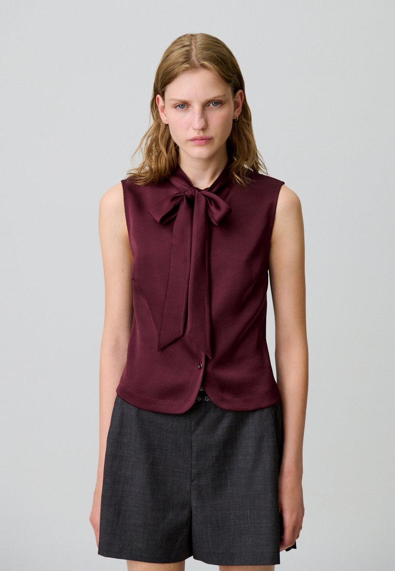 Blusa senza maniche bordeaux con scollatura con nodo, tessuto liscio e chiusura a bottoni nascosta, abbinata a pantaloni bermuda grigi scuri.