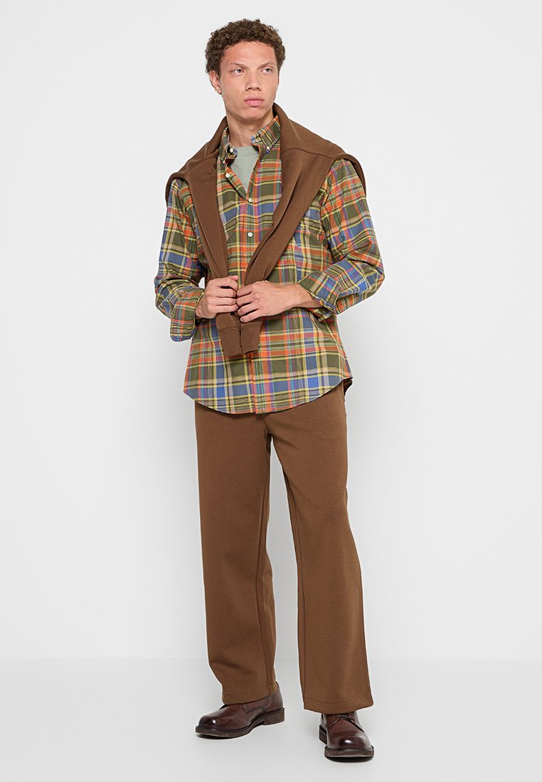 Un pull brun drapé sur les épaules, une chemise à carreaux multicolore avec des nuances de vert, rouge et bleu, assortie à un pantalon large brun et des chaussures brunes.