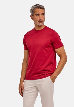 Uomo con capelli grigi e barba che indossa una camicia rossa a maniche corte e pantaloni beige chiaro, in piedi con una mano in tasca su sfondo bianco.