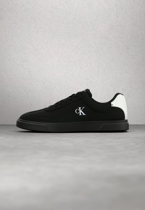 Schwarzer Low-Top-Sneaker mit weißem Fersenpatch und weißem "cK" Logo an der Seite, auf grauer Oberfläche und Hintergrund präsentiert.