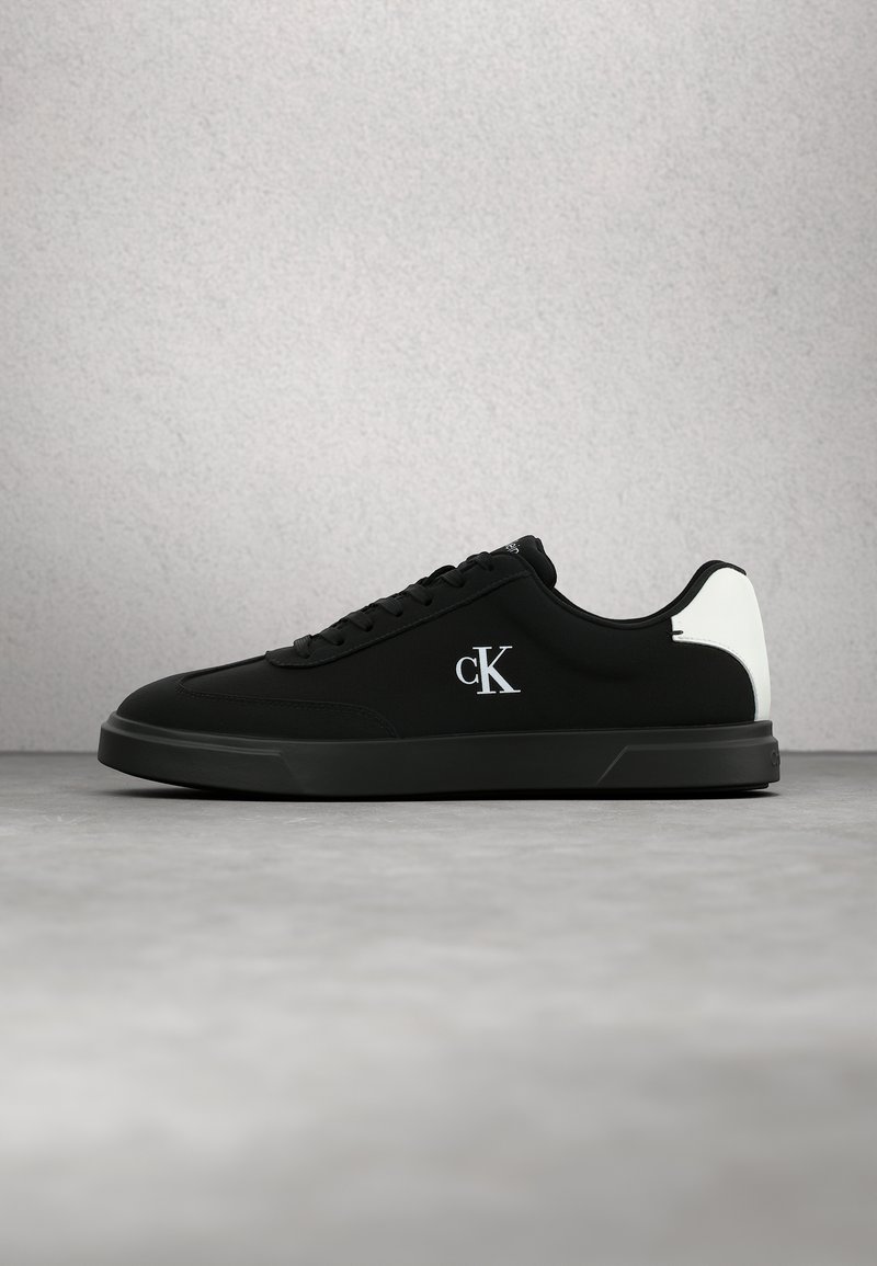 Baskets basses noires avec un patch blanc au talon et un logo blanc "cK" sur le côté, présentées sur une surface et un fond gris.