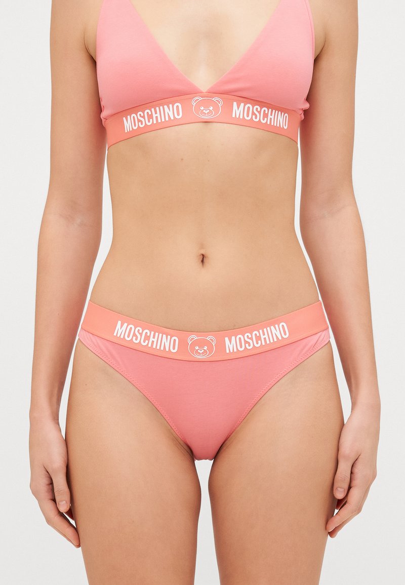 Rozā kokvilnas bralete un atbilstošas biksītes ar lāča logotipu un balto "MOSCHINO" tekstu uz jostas, ar vienkāršu, gludu dizainu.