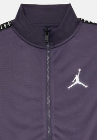Mørk lilla sportsjakke med hel glidelås og hvit Jumpman-logo på venstre bryst, svart skulderbånd med hvit "JORDAN" tekst.