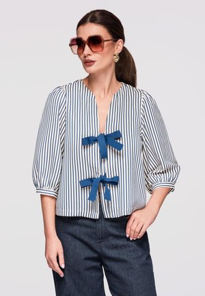 Femme portant un chemisier à rayures verticales bleu et blanc avec deux nœuds bleus à l'avant, pantalon sombre, lunettes de soleil surdimensionnées et boucles d'oreilles en forme de cœur.