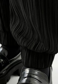 Schwarze plissierte Hosen aus strukturiertem Stoff, kombiniert mit polierten schwarzen Loafers mit horizontalem Ausschnittdesign, die auf einer schwarzen Socke ruhen.