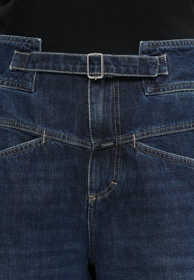 Tumši zili denim džinsi ar jostu, kurai ir taisnstūra sudraba sprādze. Redzama šuvju apdare un uzlieka etiķete ar uzrakstu "CLOSED" netālu no jostasvietas.