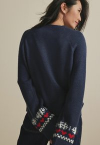 Pull bleu marine avec une finition texturée. Présente des cœurs rouges à motifs et des flocons de neige blancs aux poignets. Col rond, coupe décontractée.