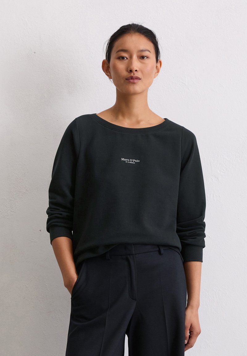 Felpa verde scuro con scollatura ampia, maniche lunghe e un piccolo logo bianco sul petto. Abbinata a pantaloni blu navy.