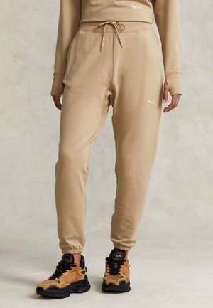 Persoon draagt beige joggerbroek met trekkoord en bijpassend top, gecombineerd met beige en zwarte Polo sneakers op een lichte achtergrond.