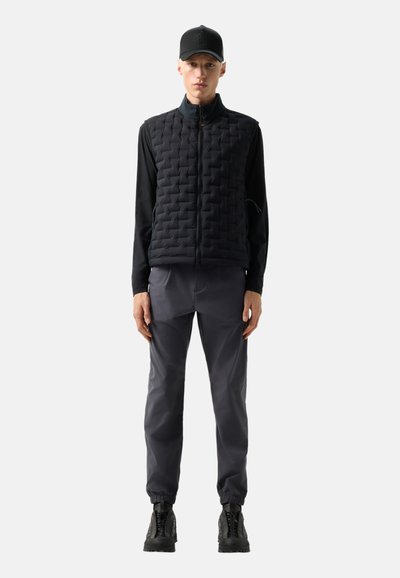 Ein Mann steht aufrecht und trägt eine schwarze gesteppte Weste, ein langärmeliges schwarzes Hemd, dunkelgraue Hosen mit elastischen Bündchen, eine schwarze Mütze und schwarze Wanderschuhe.