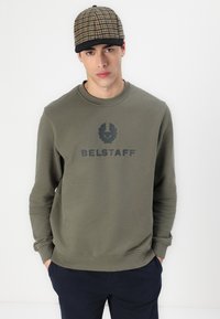 Maslinasto zeleni dukserica s okruglim izrezom i rebrastim rukavima, s istaknutim crnim logom i natpisom. Model nosi šešir s karo uzorkom.