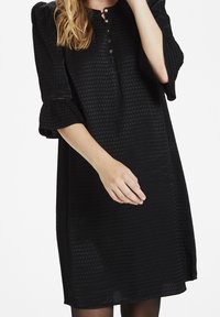 Svart klänning med texturerad  tyg, knappslå och utsvängt ärm. Mjuk textur med en fåll ovanför knät.