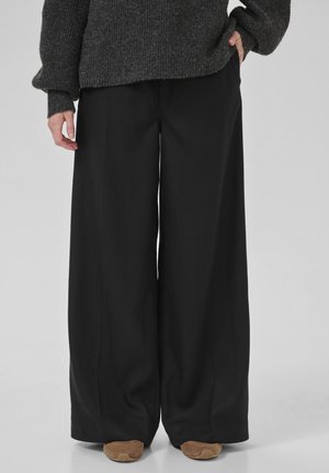 Person trägt locker sitzende schwarze Hosen mit weitem Bein, braune Schuhe und einen dunkelgrauen Strickpullover, steht mit einer Hand in der Tasche.