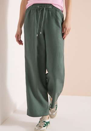 Personne portant un pantalon vert à jambes larges avec un cordon de serrage et des baskets beiges avec des accents verts, debout devant un fond pâle.