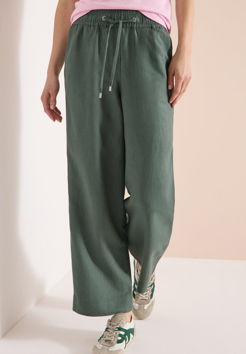 Personne portant un pantalon vert à jambes larges avec un cordon de serrage et des baskets beiges avec des accents verts, debout devant un fond pâle.