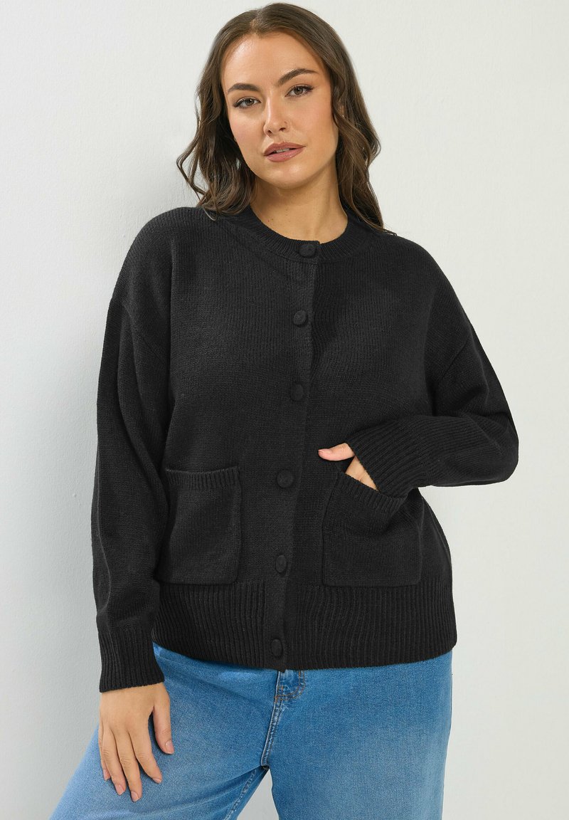 Frau mit offenem braunem Haar, die eine schwarze, mit Knöpfen versehene Strickjacke mit zwei Vordertaschen und blaue Jeans trägt, stehend vor einer schlichten weißen Wand.