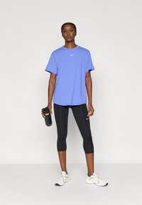 Μωβ κοντομάνικο μπλουζάκι Nike συνδυασμένο με μαύρο κολάν κάπρι. Το μοντέλο κρατάει ένα μαύρο παγούρι και φοράει λευκά αθλητικά παπούτσια.