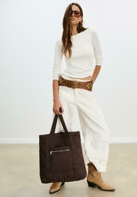 Borsa tote in tessuto marrone con motivo trapuntato e tasca frontale con zip, di dimensioni medie, tenuta da un modello che indossa una maglietta bianca a maniche lunghe e jeans a gamba larga.