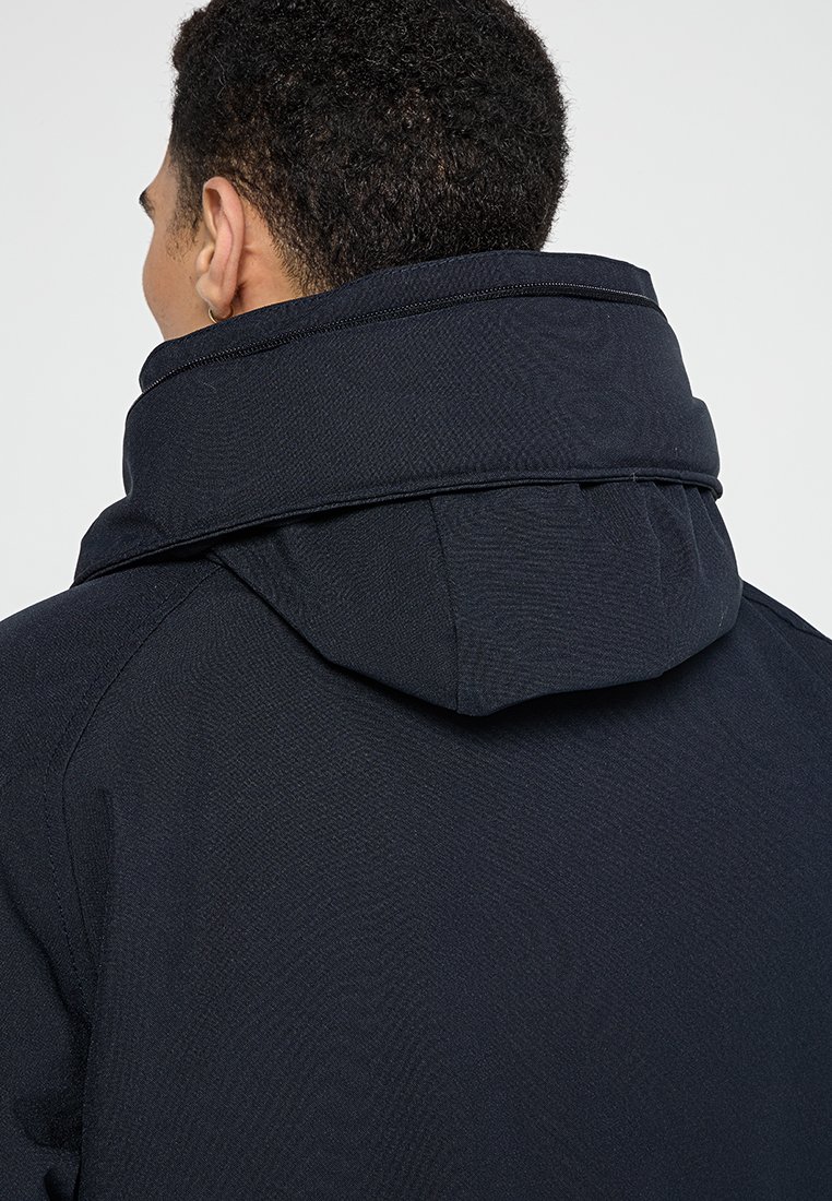 Veste navy avec un col haut et une capuche. Le tissu présente un motif texturé et des coutures renforcées le long des seams.