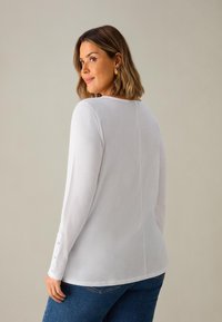 Camicia bianca a maniche lunghe con scollatura rotonda, con dettagli a bottoni sulle maniche. Il materiale appare morbido con una texture liscia.