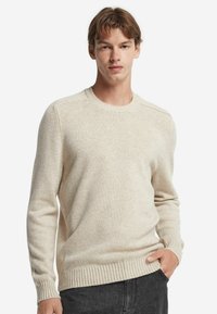 Beżowy sweter dzianinowy z okrągłym dekoltem, ściągaczami przy mankietach i na dole. Charakteryzuje się gładką fakturą i luźnym krojem, noszony na ciemnoszarych spodniach.