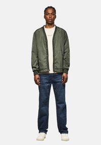 Olivgrüne Bomberjacke mit Knopfverschluss und gerippten Bündchen, kombiniert mit einem hellbeigen Hemd und dunkelblauen Jeans. Weiße Sneaker.