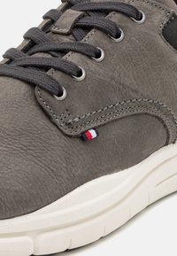 Šedá kožená sneakers s texturovaným povrchem, kulatou špičkou, plochými tkaničkami, kovovými očky a malou trojbarevnou nášivkou poblíž tkaniček.