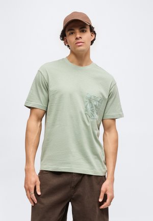 Giovane uomo che indossa una maglietta a righe verde chiaro con tasca sul petto decorata, pantaloni marroni, cappellino beige e anelli d'argento, in piedi davanti a uno sfondo grigio.