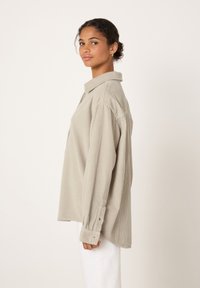 Gaiši bēša korduroja krekls ar apkakli, garām piedurknēm un pogu aprocēm. Raksturots ar brīvu piegriezumu un teksturētu audumu.