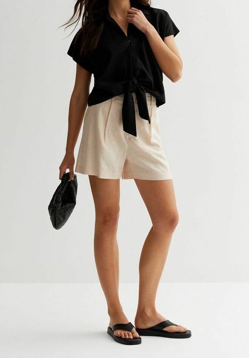 Chemise noire à manches courtes avec lien noué à l'avant, short beige plissé et sandales plates noires. Tenant une pochette noire froissée.
