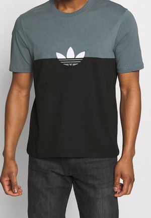 Mann trägt ein kurzärmeliges T-Shirt mit Rundhalsausschnitt, grauer oberer Hälfte, schwarzer unterer Hälfte und weißem Adidas-Trefoil-Logo auf der Brust.