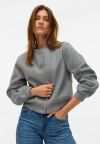 Chaqueta bomber gris con cuello redondo, botones frontales y puños acanalados. Hecha de un material suave y texturizado, combinada con jeans de mezclilla azul.