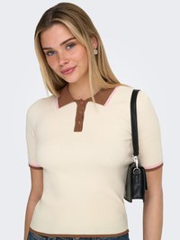 Mladi žena s ravnom plavom kosom nosi kremasti rebrasti top s kratkim rukavima s smeđim ovratnikom i manžetama, nosi crnu torbu preko ramena.