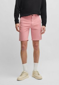 Pantalones cortos de algodón rosa con un frente plano, bolsillos laterales y detalle en el bolsillo trasero. Combinados con un sudadera negra y zapatillas beige.
