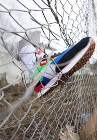 Baskets avec une tige noire, bleue et verte, des détails blancs, des accents rouges et une semelle en caoutchouc texturée, reposant sur une clôture en mailles métalliques.