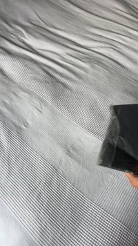 Čierny predmet v ruke nad sivou posteľnou prikrývkou s textúrou s horizontálnymi čiarami a vzorovanými časťami. Mäkký, hladký materiál s rôznymi vzormi.