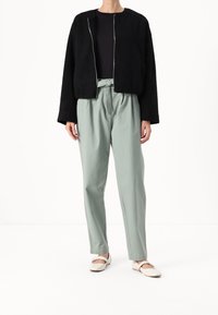 Giacca in pile nera con zip argento, abbinata a pantaloni verde chiaro a vita alta e ballerine beige. Design semplice e vestibilità comoda.